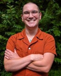 A professional photo of Kai wearing a dark orange jumpsuit. Their arms are crossed in front of them and they have a big smile.